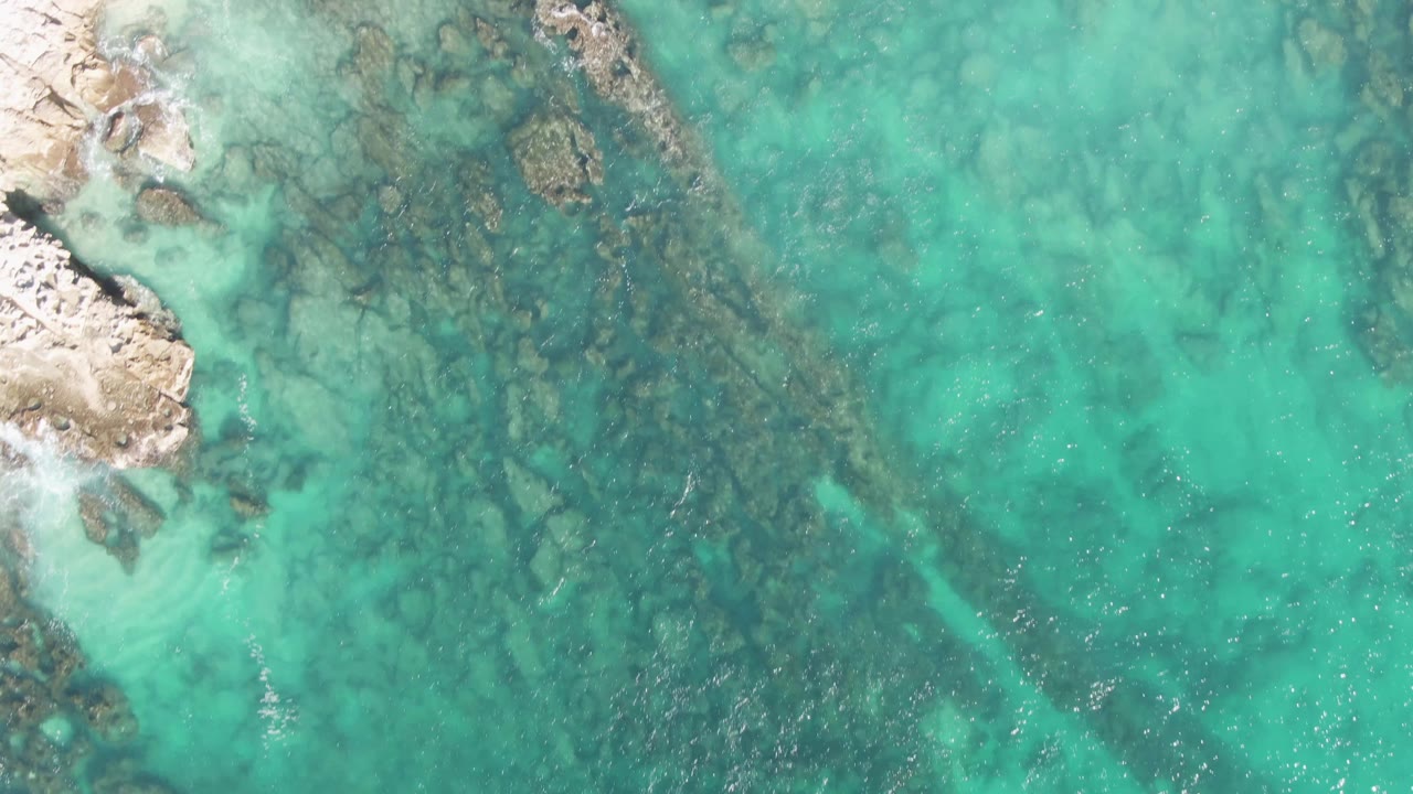 Straight-down aerial view at Buzzards Beach, Baja California Sur, Mexico, showing turquoise water over dark submerged rocks with pale shore rock at the left edge and no visible people.