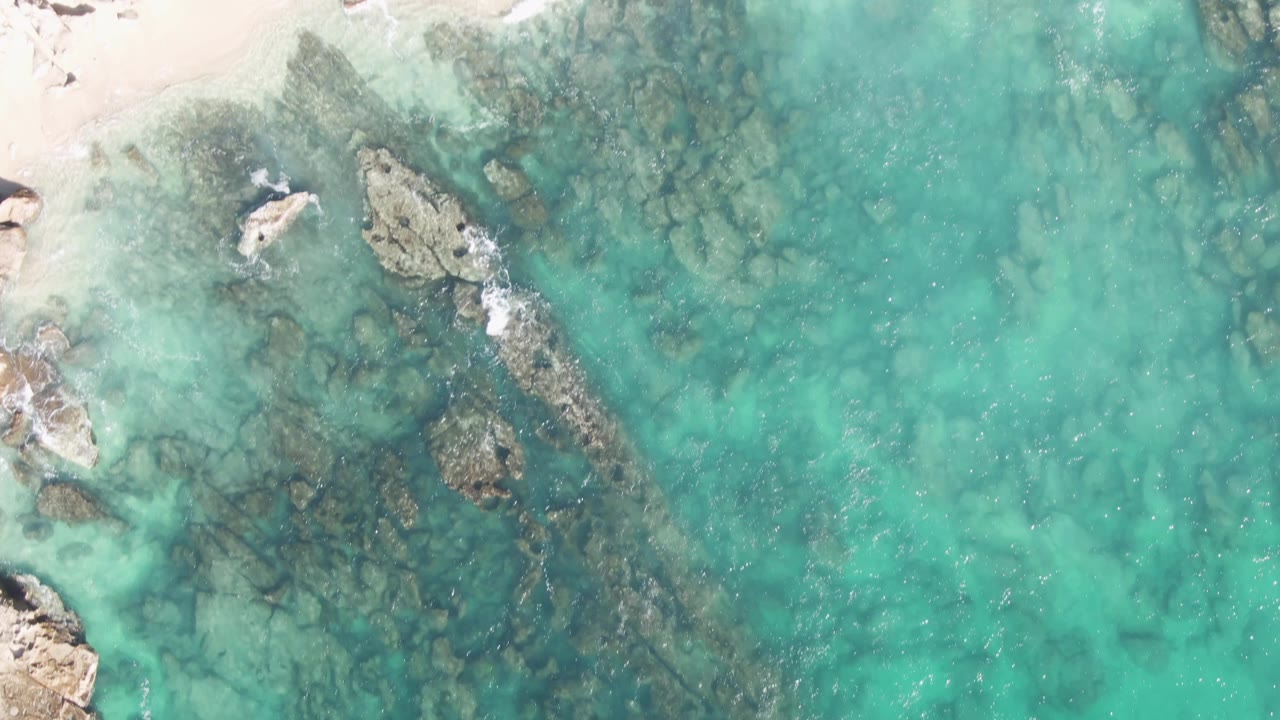 Buzzards Beach aerial view with pale sand entering the top left, turquoise water over submerged rocks, small exposed rocks, and no visible people in the frame.