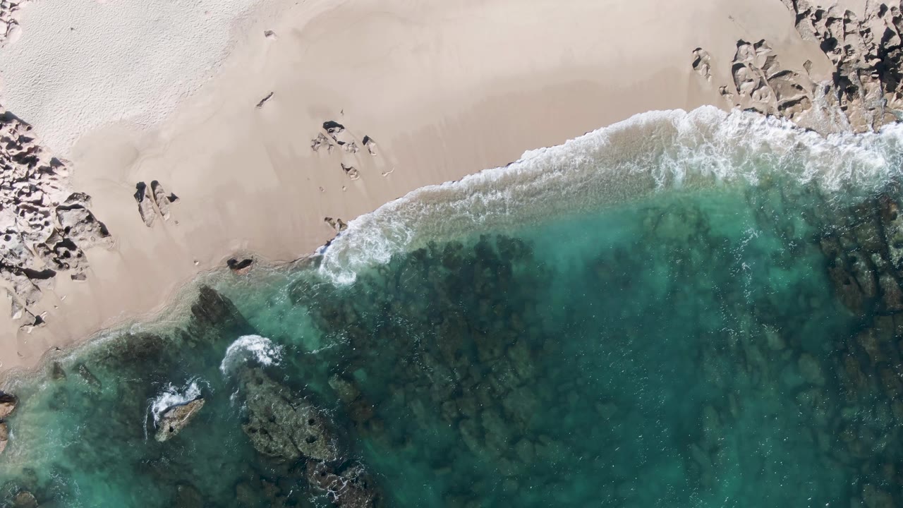 Overhead frame of Buzzards Beach in Baja California Sur showing two small people on pale sand above turquoise water, with white surf and dark rocks along the shore.