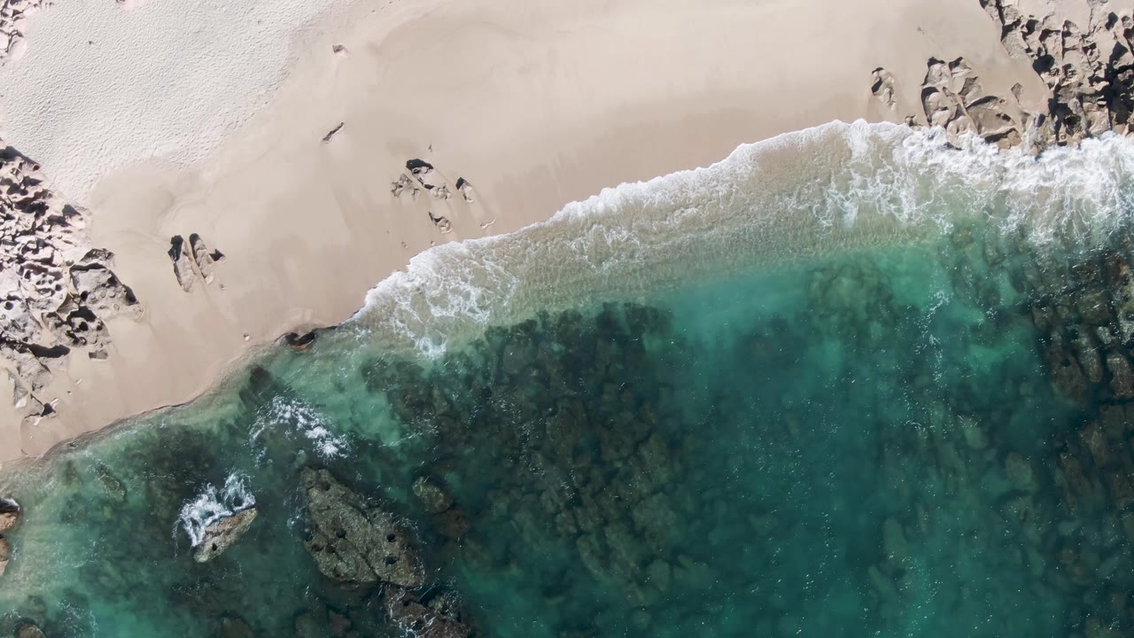 Aerial shoreline with walkers on sand, foamy wave edge, clear water, and underwater rocks.