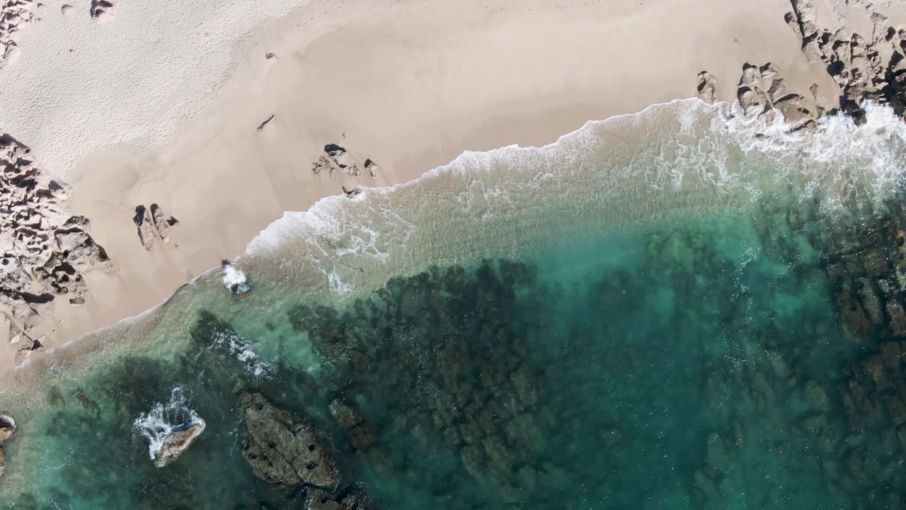 At 0:02, the Buzzards Beach overhead view places the two people near the upper sand with a softer foam band along the shoreline and dark submerged rock patches below.