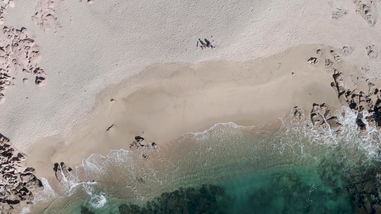 Top-down view of Buzzards Beach in Baja California Sur with pale sand, dark rocks, turquoise water, thin foam, and small people visible near the shoreline.