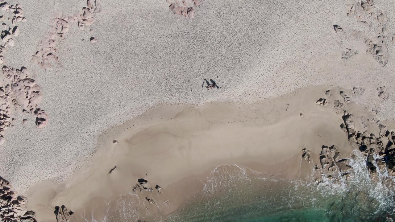 Top-down Buzzards Beach frame with broad pale sand, a curved wet shoreline, rocks along the right side, turquoise water at the bottom, and small people visible on the sand.