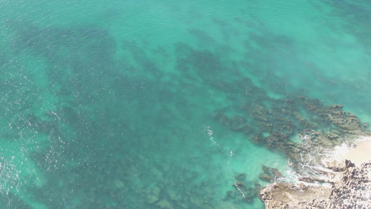 Aerial frame at Buzzards Beach in Baja California Sur, Mexico, with darker underwater rock patches beneath turquoise water and a small foamy break along the lower-right rocks, no visible people.