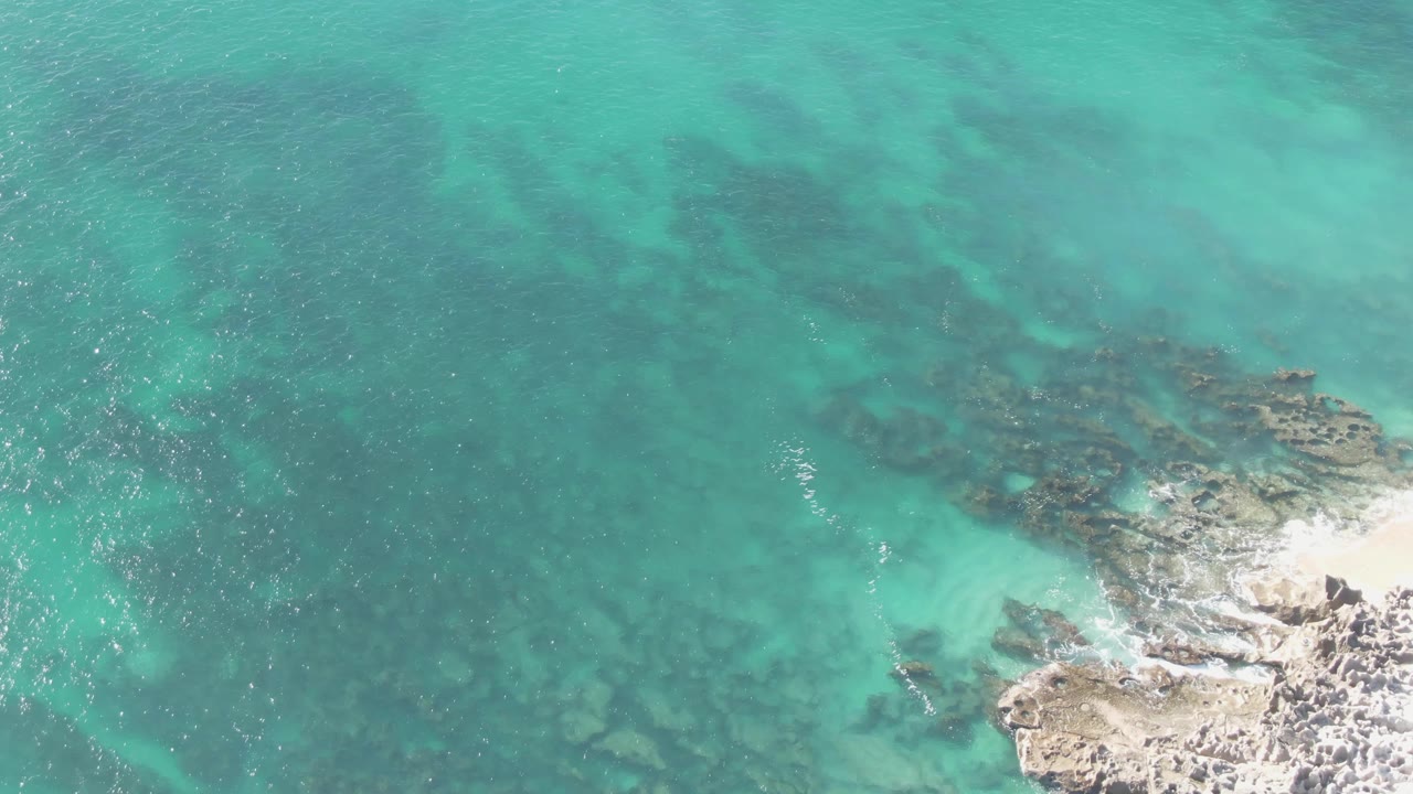 Top-down view of blue-green water at Buzzards Beach in Baja California Sur, Mexico, with pale rocks at the lower right and a thin line of white wash, no visible people.