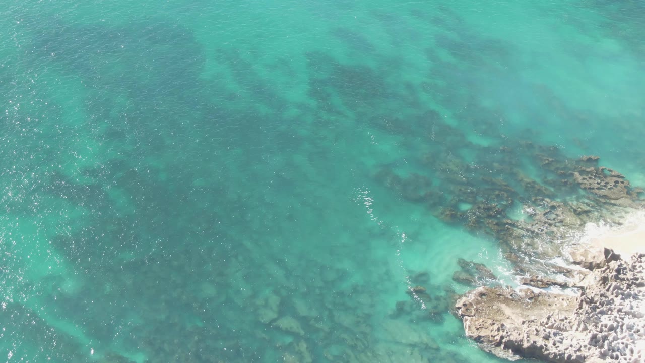 Mid-clip aerial view at Buzzards Beach in Baja California Sur, Mexico, where translucent turquoise water covers submerged rocks and foam gathers against the lower-right shore, no visible people.