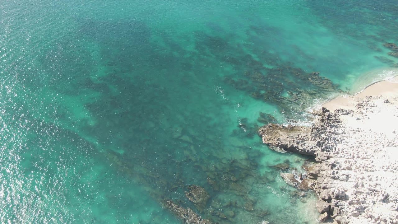 A rocky point at Buzzards Beach projects into turquoise shallows, with pale sand near the upper right and darker reef-like patches visible below the surface, no visible people.