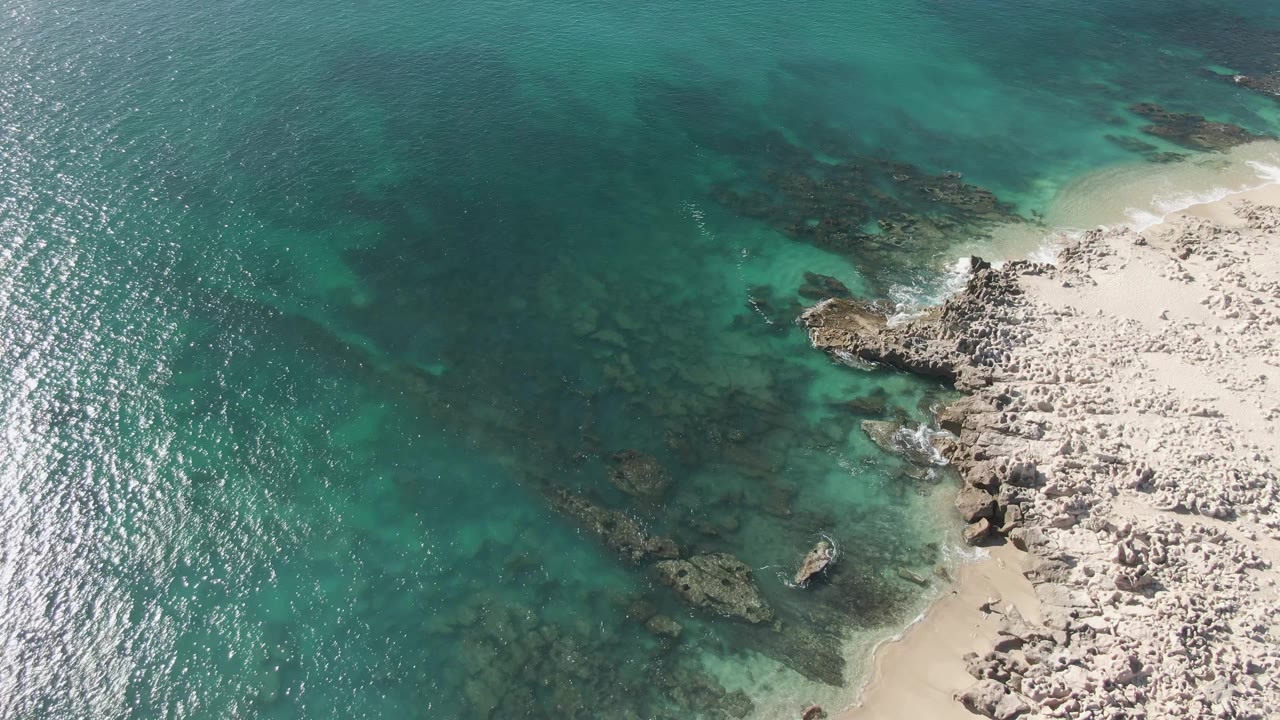 Aerial view of turquoise water, submerged rocks, sandy shore, and white foam.