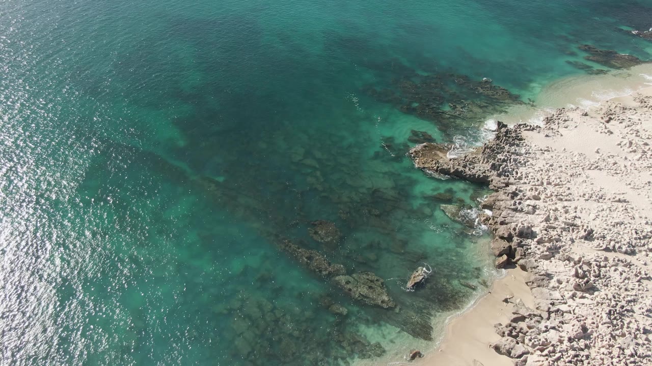 Aerial view at Buzzards Beach with the rocky sand edge on the right, darker seabed shapes under green-blue water, small foam lines, and no visible people.