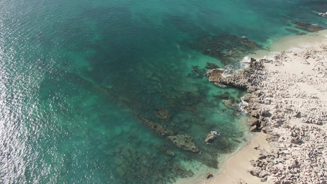 Aerial Buzzards Beach frame with the coastline pushed to the right, dark reef-like rock patches visible below turquoise water, bright shore rocks, and no visible people.