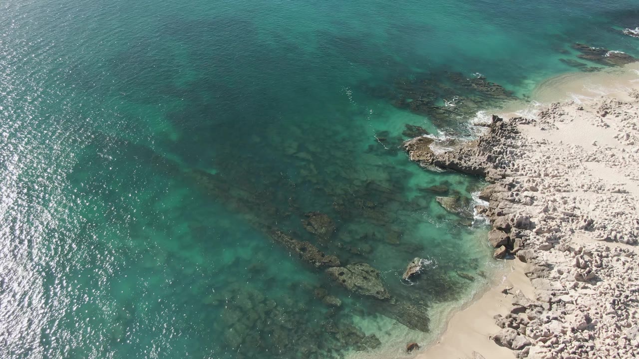 Aerial view over Buzzards Beach showing a wider band of sun sparkle on rippled water, pale sand below the rocky edge, submerged rocks, and no visible people.