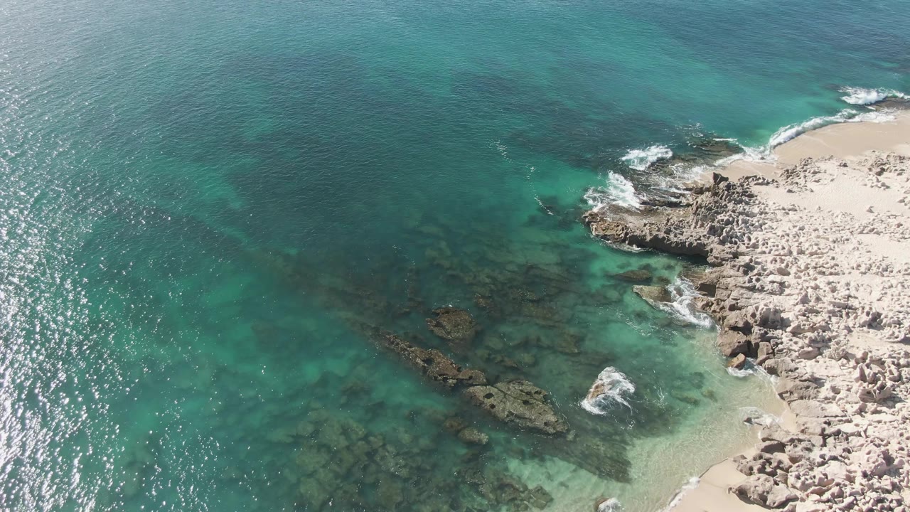 Buzzards Beach aerial frame with clear turquoise water covering dark underwater rocks, a foamy break near the shoreline, pale sand, and no visible people.