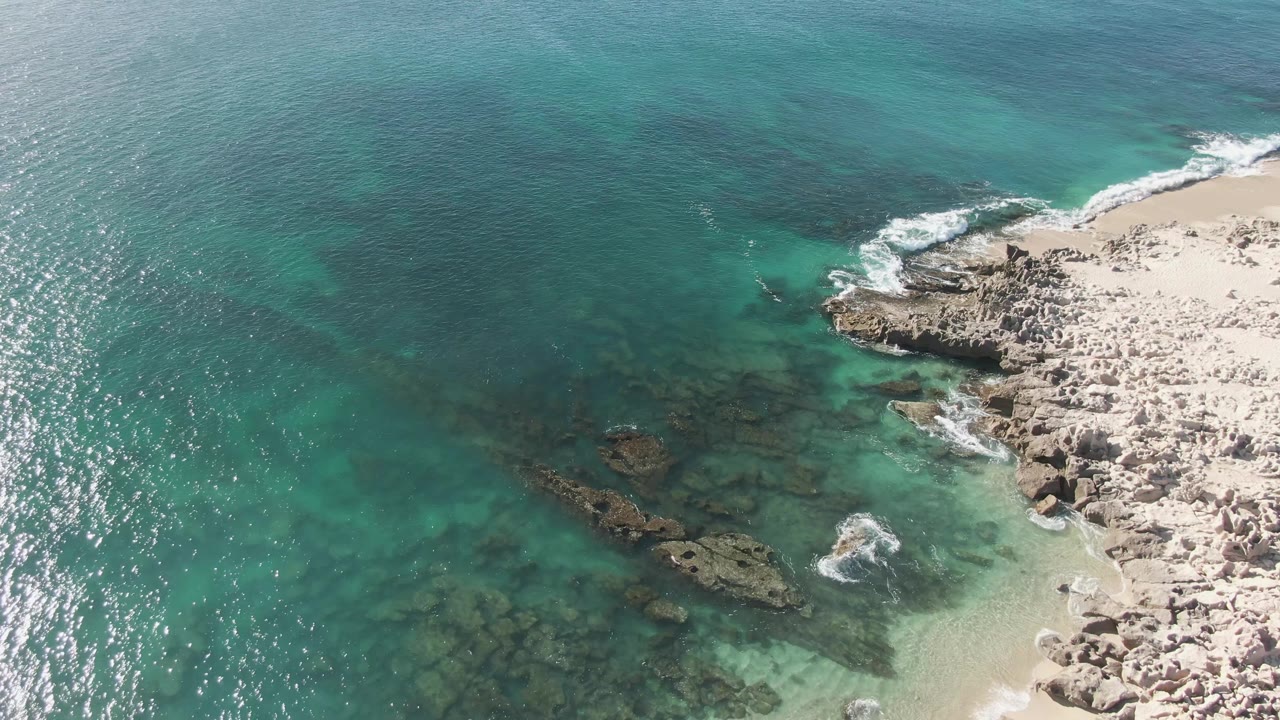 High aerial view of the Buzzards Beach shoreline with blue-green ocean, exposed rocks, submerged reef-like shapes, thin foam lines, and no visible people.