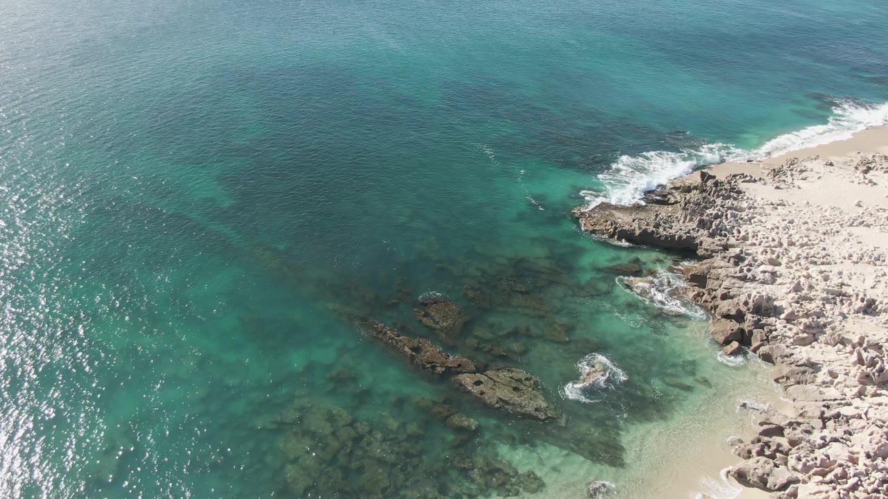 Drone-style view over Buzzards Beach with turquoise water occupying most of the frame, dark rock patches below the surface, pale shoreline rocks, and no visible people.