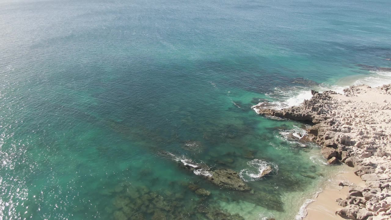 Aerial frame over Buzzards Beach with clear green-blue water exposing dark reef shapes, a pale rocky coast at right, small white breakers, and no visible people.