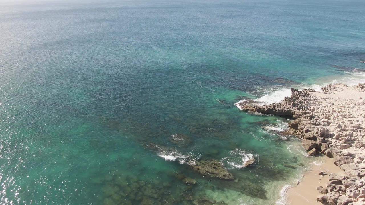 Aerial view at Buzzards Beach showing more open blue ocean in the upper frame, turquoise shallows over rocks below, surf curling near the right shoreline, and no visible people.
