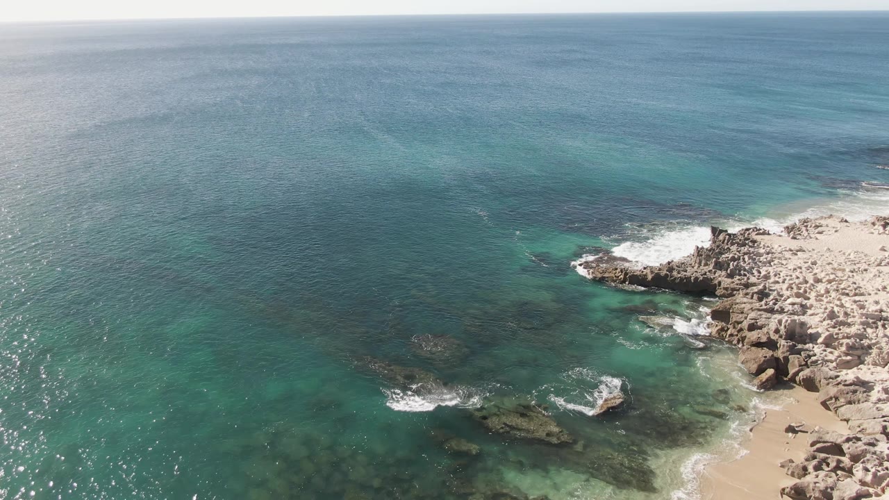 Aerial frame over Buzzards Beach where sun flecks mark the left water surface, dark submerged rocks sit in the center, waves touch the right-side rocks, and no visible people appear.
