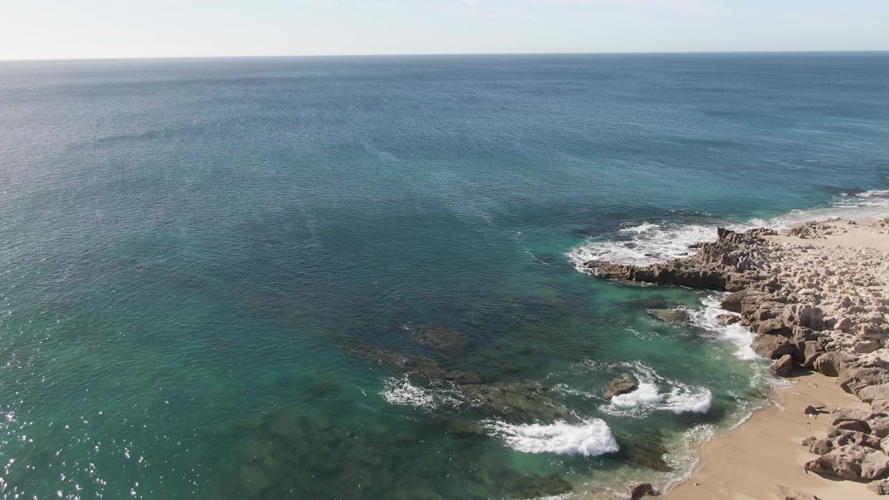 Aerial view of turquoise ocean, rocky shoreline, sand, and white surf.