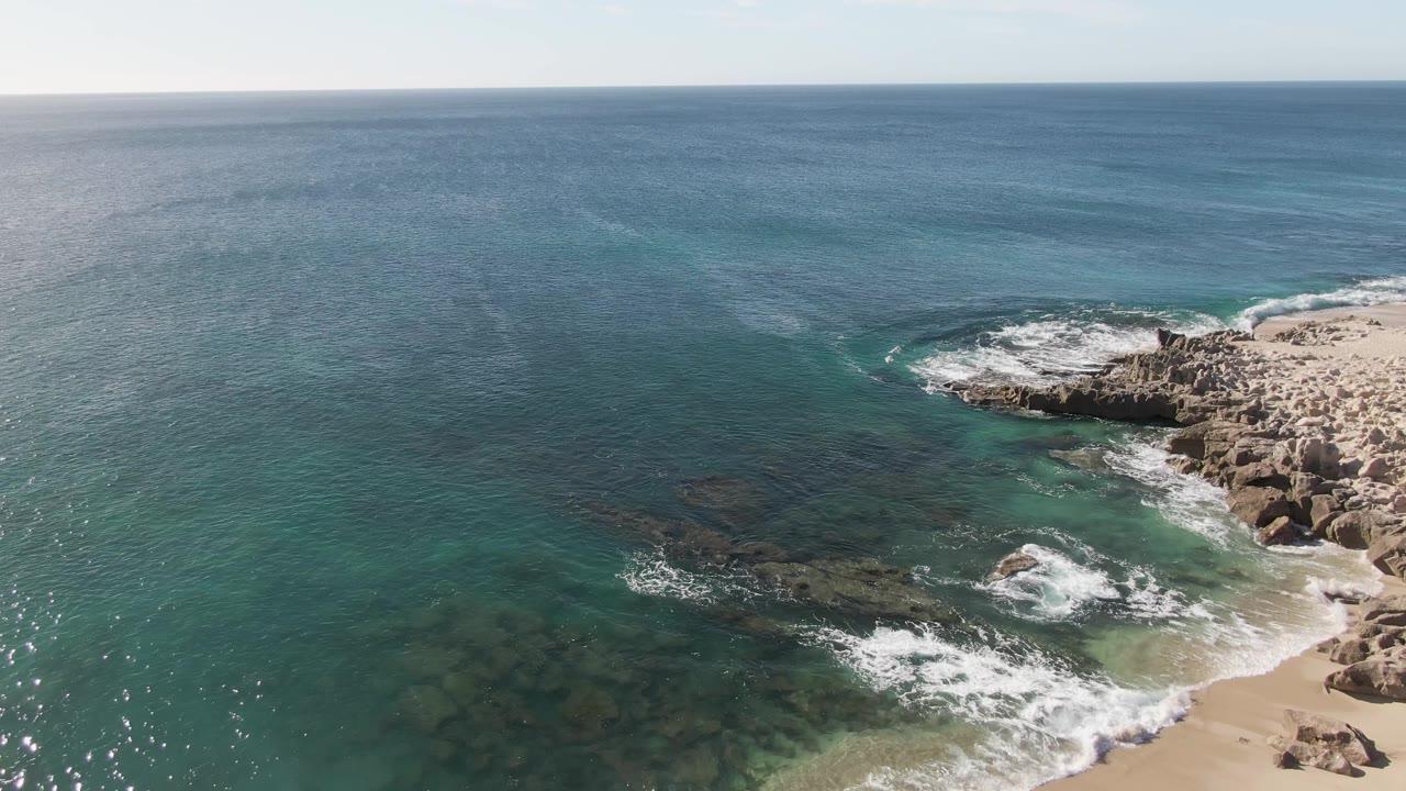 Aerial view at Buzzards Beach with a rocky point on the right, white surf curling around the rocks, shallow green-blue water below, and no visible people.