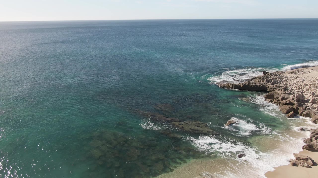 Wide aerial frame at Buzzards Beach with more open ocean filling the left side, rocky coastline along the right, foam over shallow rocks near shore, and no visible people.