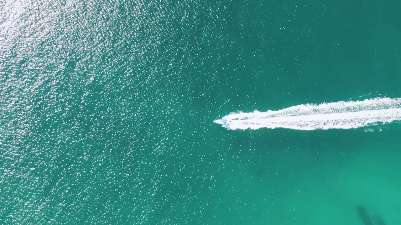 At Buzzards Beach in Baja California Sur, the boat sits farther across the frame as its white foamy wake widens over sunlit turquoise sea, with no visible people.