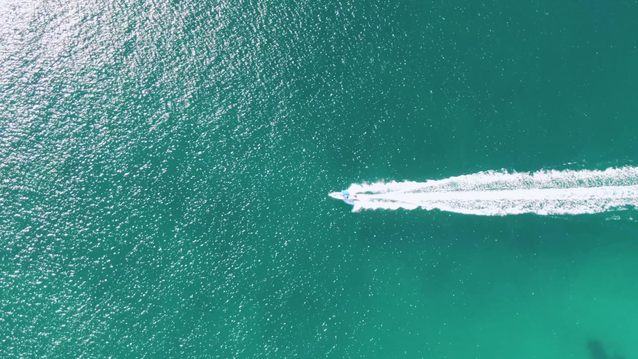At Buzzards Beach in Baja California Sur, the speedboat approaches the right side of the overhead frame, pulling a long white wake through open turquoise water, with no visible people.