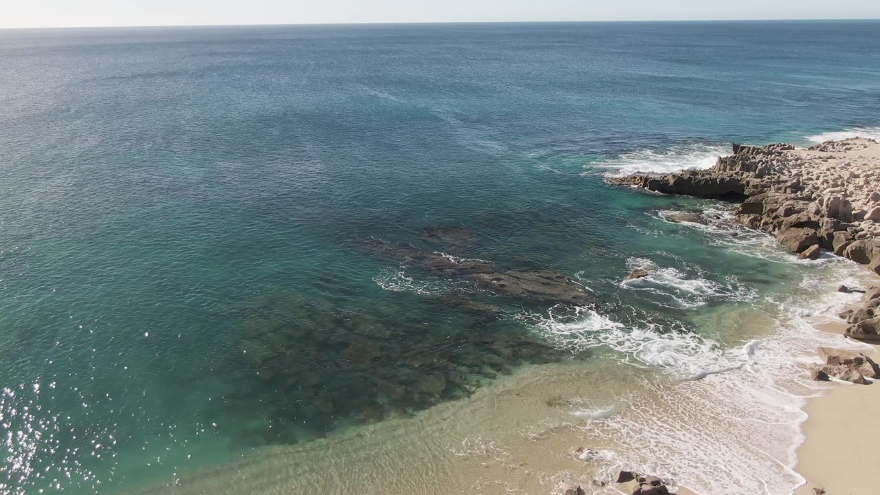 Aerial view at Buzzards Beach, Baja California Sur, showing turquoise water, white foam, sandy shore, rocky outcrop, and no visible people in this opening frame.