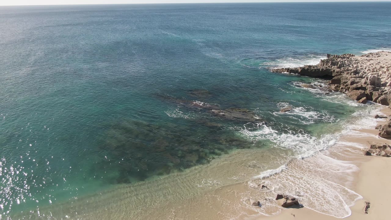 Clear ocean water and foam lines reach a sandy beach with rocks at right.