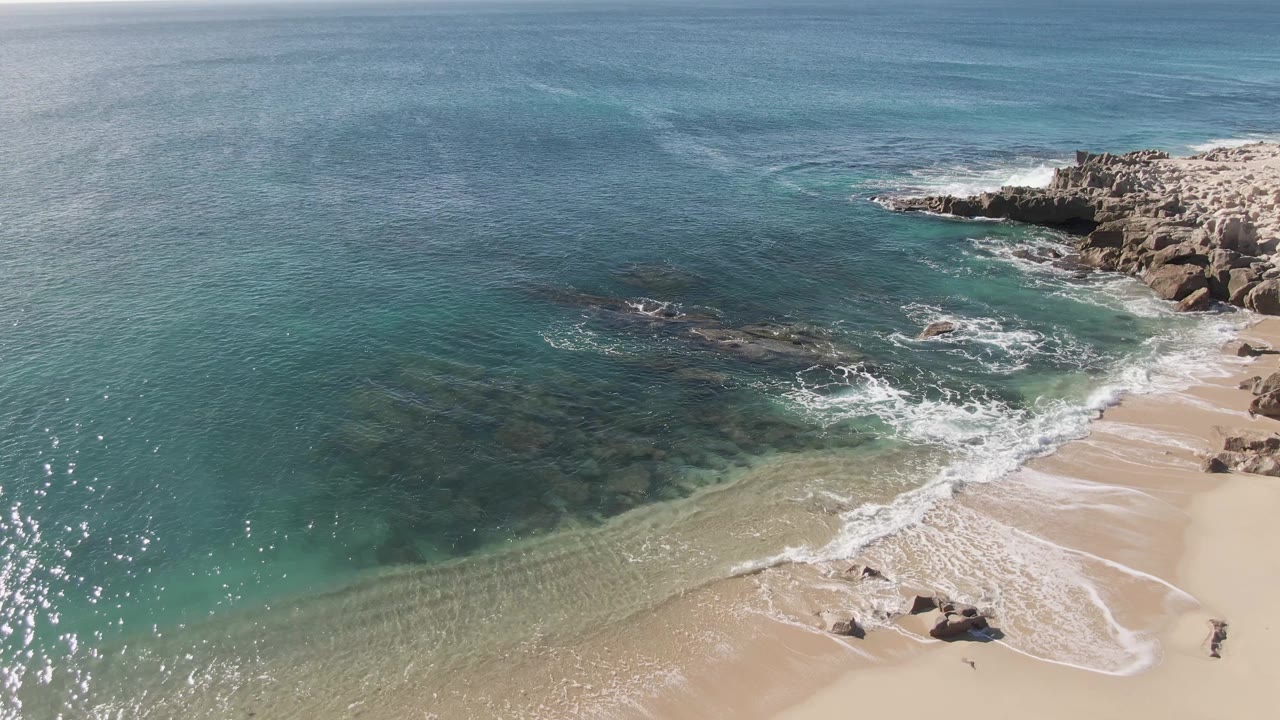 Aerial view at Buzzards Beach showing white surf curling onto sand, rocky outcrops on the right, clear water offshore, and small people walking near the lower right edge.