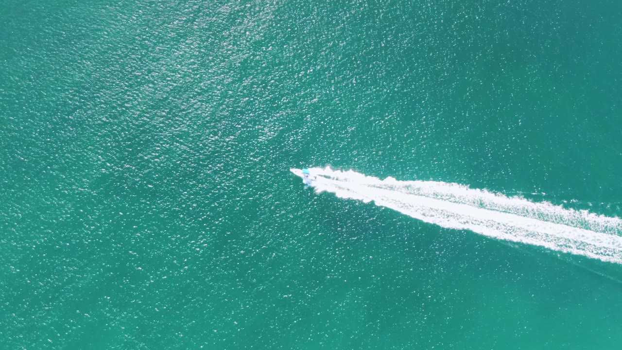 Overhead frame from Buzzards Beach, Baja California Sur, with the speedboat farther across the frame and a widening white foam trail behind it, no visible people.