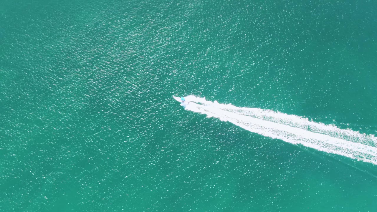 Overhead frame from Buzzards Beach, Baja California Sur, where the boat and its bright wake sit near the upper right over rippled turquoise water, no visible people.