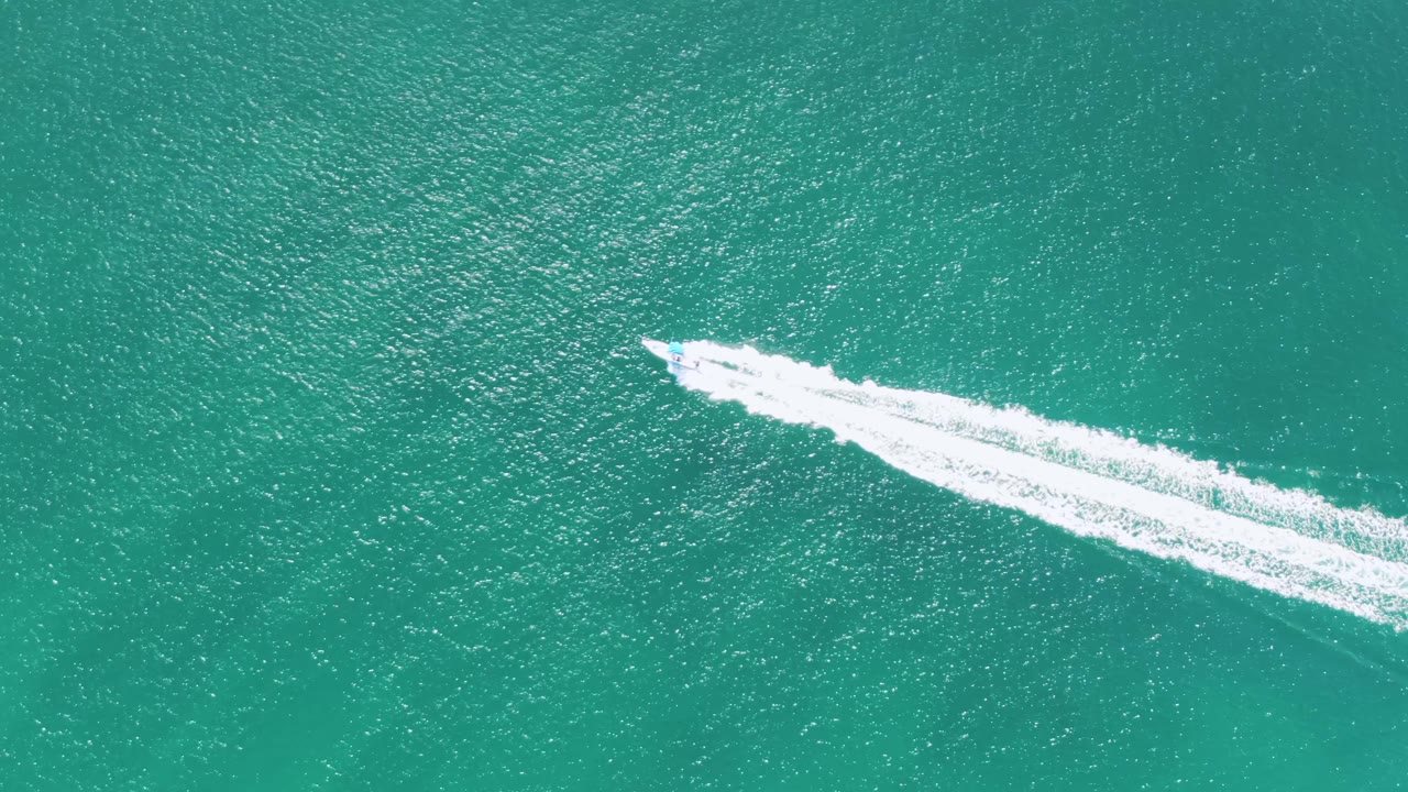 Overhead frame from Buzzards Beach, Baja California Sur, showing the boat lower in the composition with a long diagonal white wake spreading across clear turquoise water, no visible people.