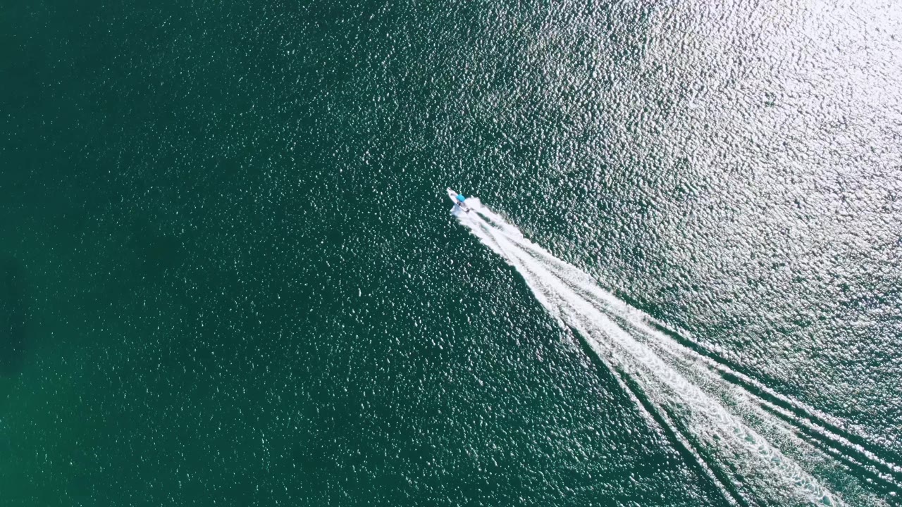 A small speedboat near the upper frame at Buzzards Beach leaves a bright white wake over dark green-blue ocean, with no shoreline or visible people in view.