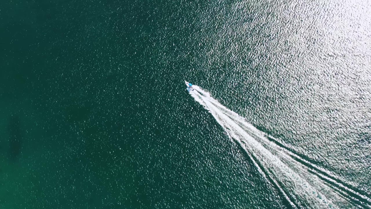 Overhead view of the Buzzards Beach ocean surface as the speedboat continues diagonally, leaving a white V-shaped wake through sunlit green-blue water and no visible people.