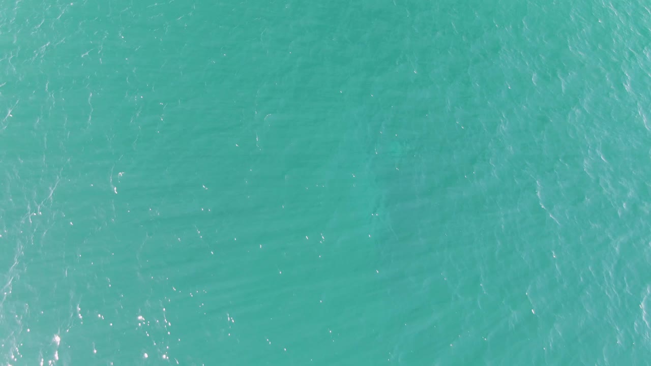 Turquoise ocean water at Buzzards Beach in Baja California Sur fills the frame as soft wave patterns cross the surface and bright flecks appear near the lower area, with no visible people.