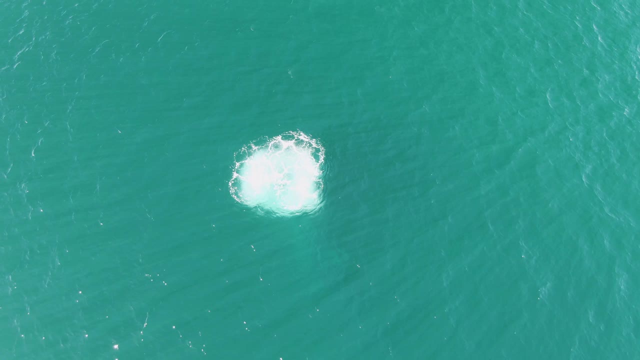 A second overhead frame at Buzzards Beach shows the white splash still bright against turquoise water, with faint surrounding ripples and no visible people.