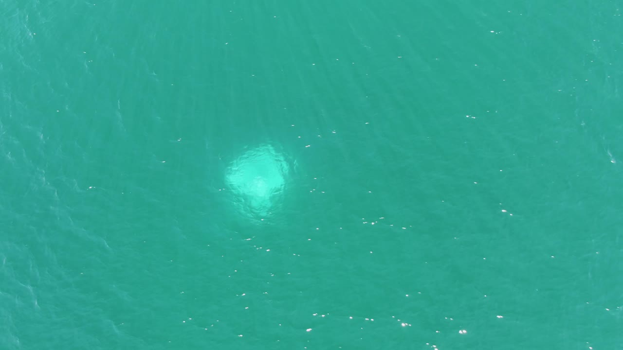 Buzzards Beach ocean surface seen from above, with a larger pale underwater light shape low in the frame, rippled turquoise water, and no visible people.