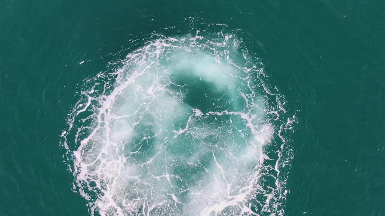 A larger white foam circle spreads across the turquoise ocean surface at Buzzards Beach, with a darker opening inside the vortex and no visible people.