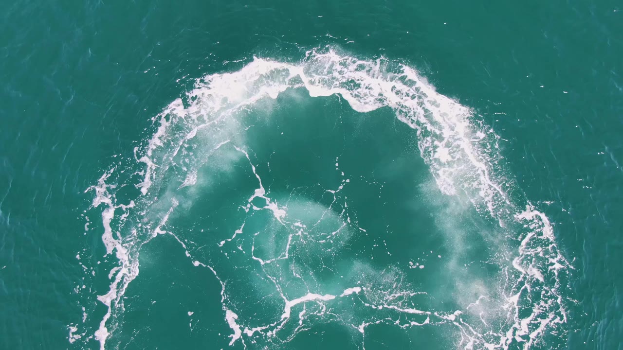 Overhead Buzzards Beach frame with the circular wake slightly more broken along the right edge as white foam spreads over turquoise ocean, no visible people.