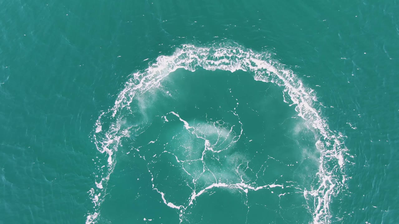 Overhead view at Buzzards Beach where the white circular wake has drifted lower in frame and is fading into softer foam on turquoise water, no visible people.