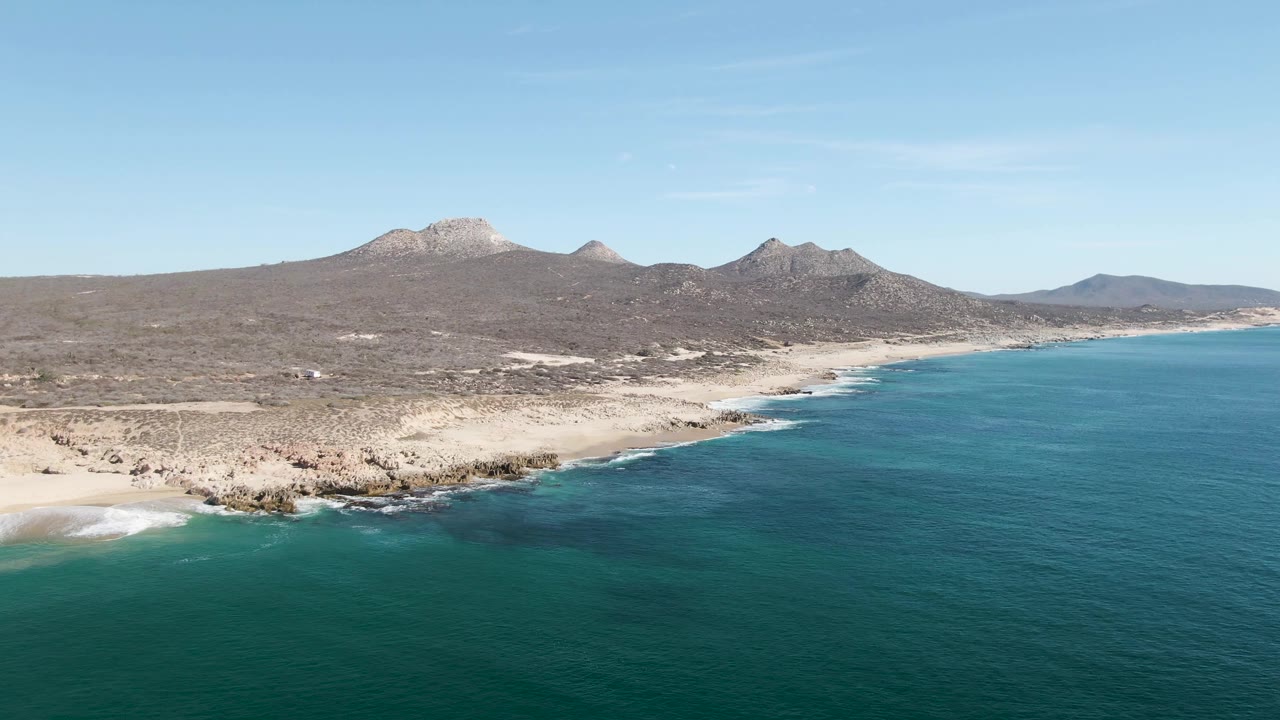 Aerial view of turquoise ocean, rocky sandy coast, and desert mountains.