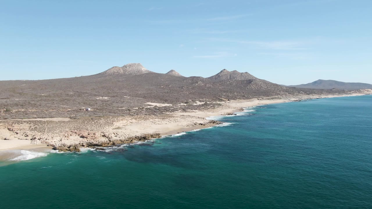 A wide Buzzards Beach aerial shows small waves breaking along the rocky edge, pale sand near the water, brown desert terrain, mountain ridges, and no visible people.