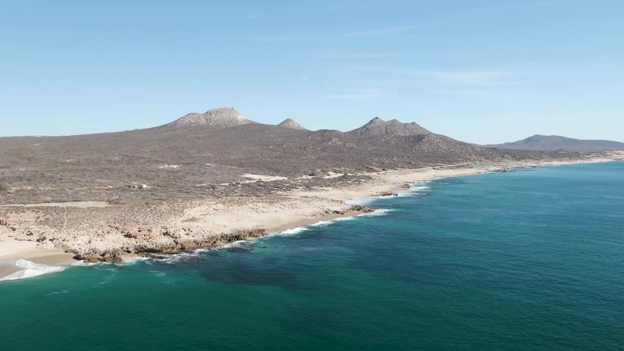The aerial view over Buzzards Beach keeps turquoise water across the foreground, a band of rocks and sand at the coast, open arid land, distant peaks, and no visible people.