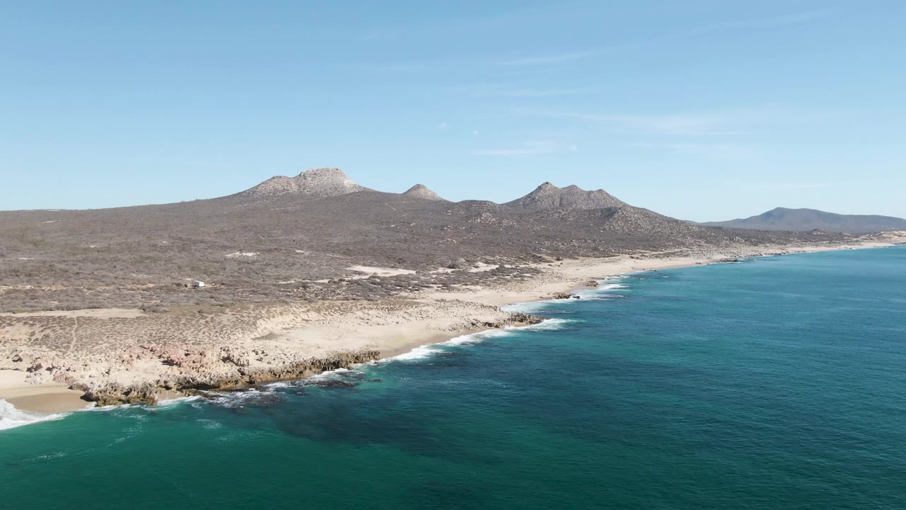 Mid-clip aerial over Buzzards Beach shows a broader sandy beach at left, dark rocks along the point, blue-green water, desert slopes, mountains, and no visible people.