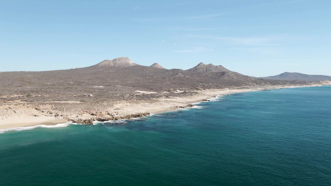 Buzzards Beach aerial frame with the shoreline angled across the scene, small white wave lines on the sand, teal water below, brown hills and mountains beyond, and no visible people.