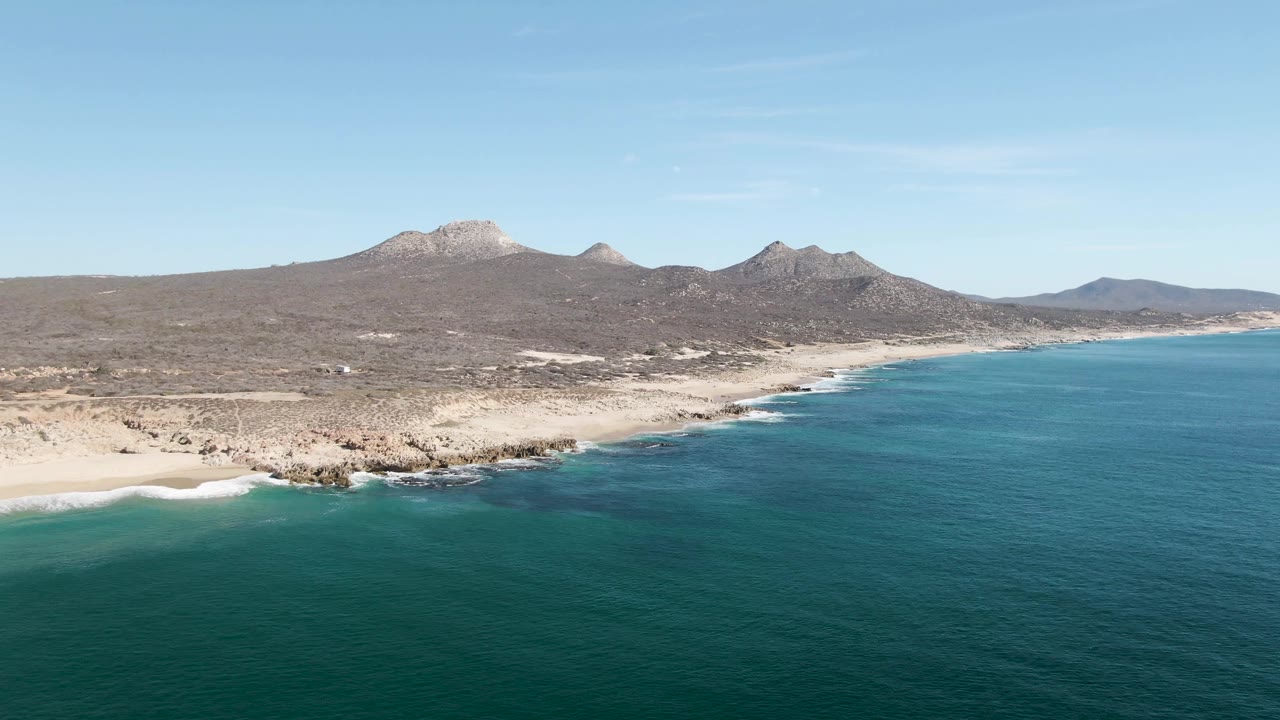Aerial view over Buzzards Beach with deeper blue-green water in the foreground, a pale beach curve at left, rocky shoreline in the center, distant mountains, and no visible people.