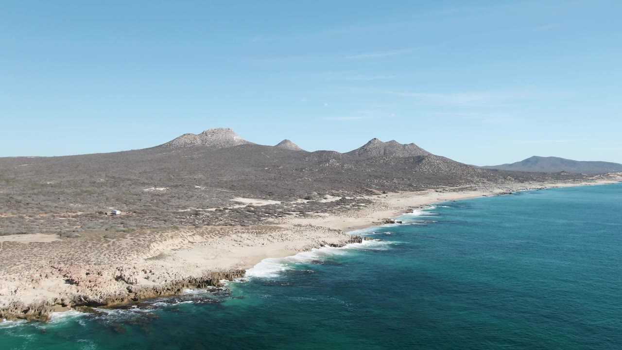 Opening aerial frame over Buzzards Beach in Baja California Sur, showing rocky shoreline at lower left, turquoise ocean at right, pale sand inland, mountain ridges on the horizon, and no visible people.