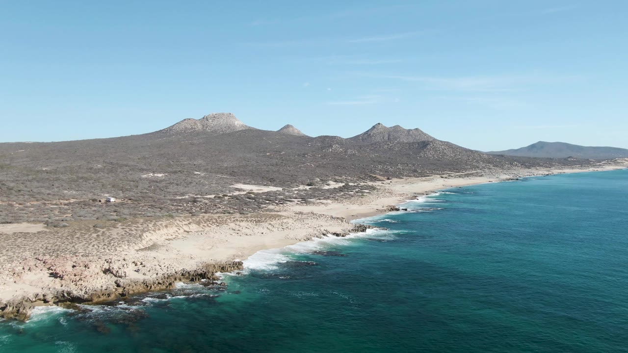 Aerial frame at Buzzards Beach showing more of the sandy shoreline curving beside turquoise water, with rocky outcrops in the lower left, dry terrain behind the beach, distant mountains, and no visible people.
