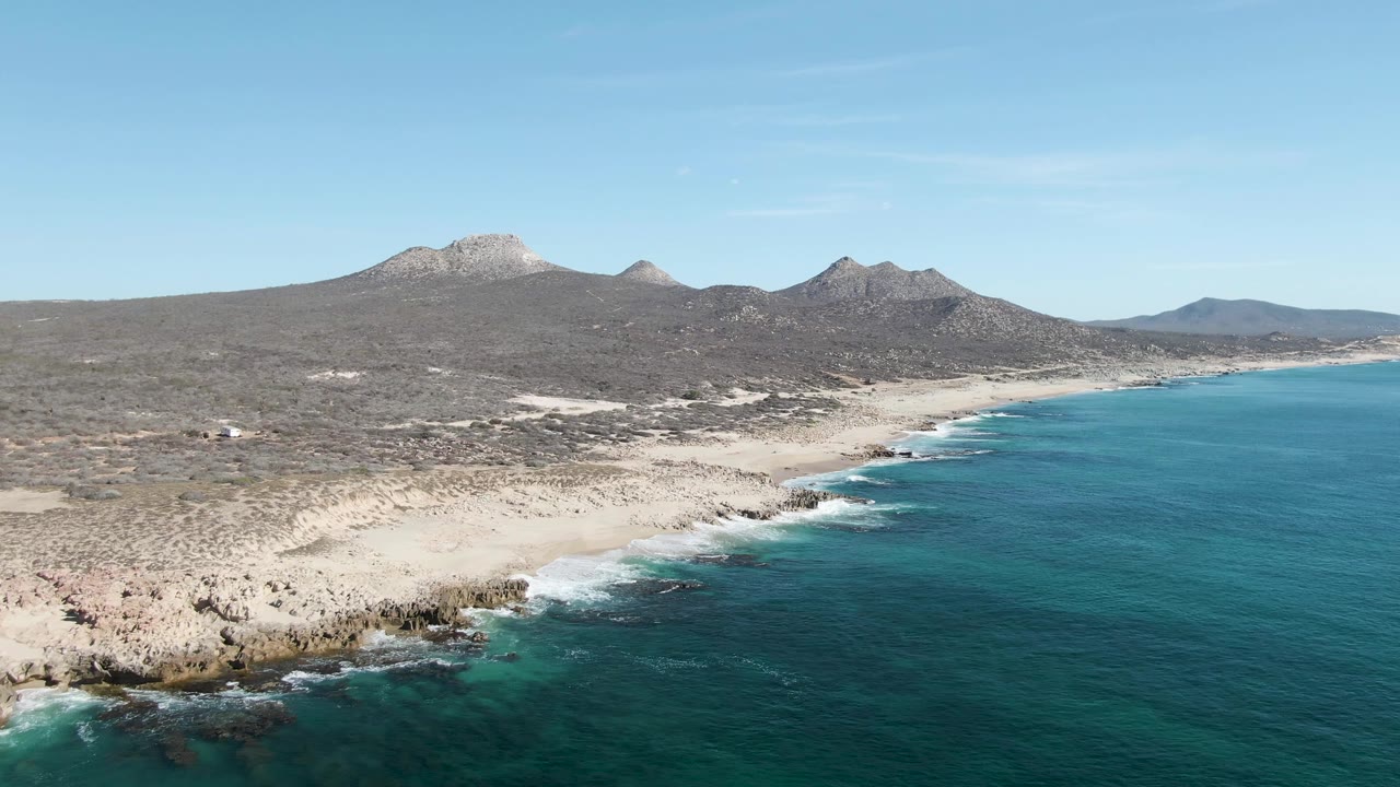 Wide aerial view of Buzzards Beach with blue-green ocean filling the right side, surf along the sand, sparse desert scrub inland, several mountain peaks across the horizon, and no visible people.