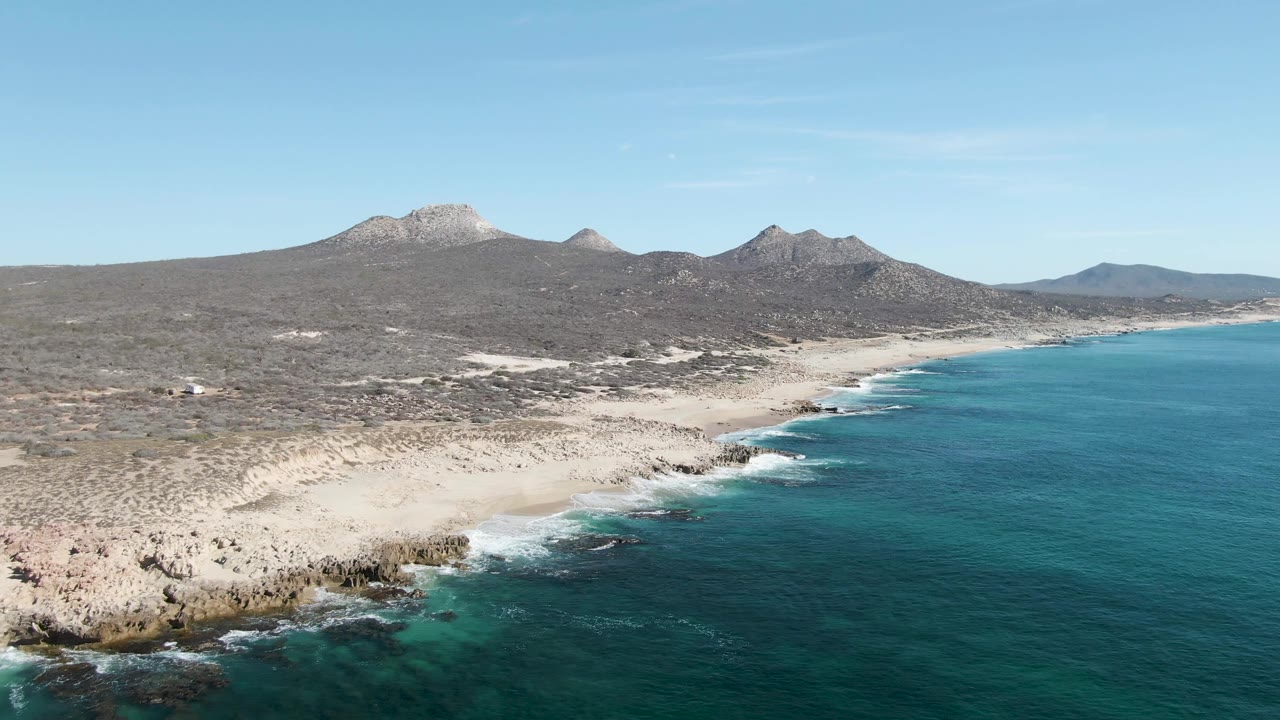 Mid-clip aerial frame over Buzzards Beach, with waves breaking against darker rocks in the lower left, a pale sandy beach stretching through the center, turquoise water to the right, mountains behind, and no visible people.
